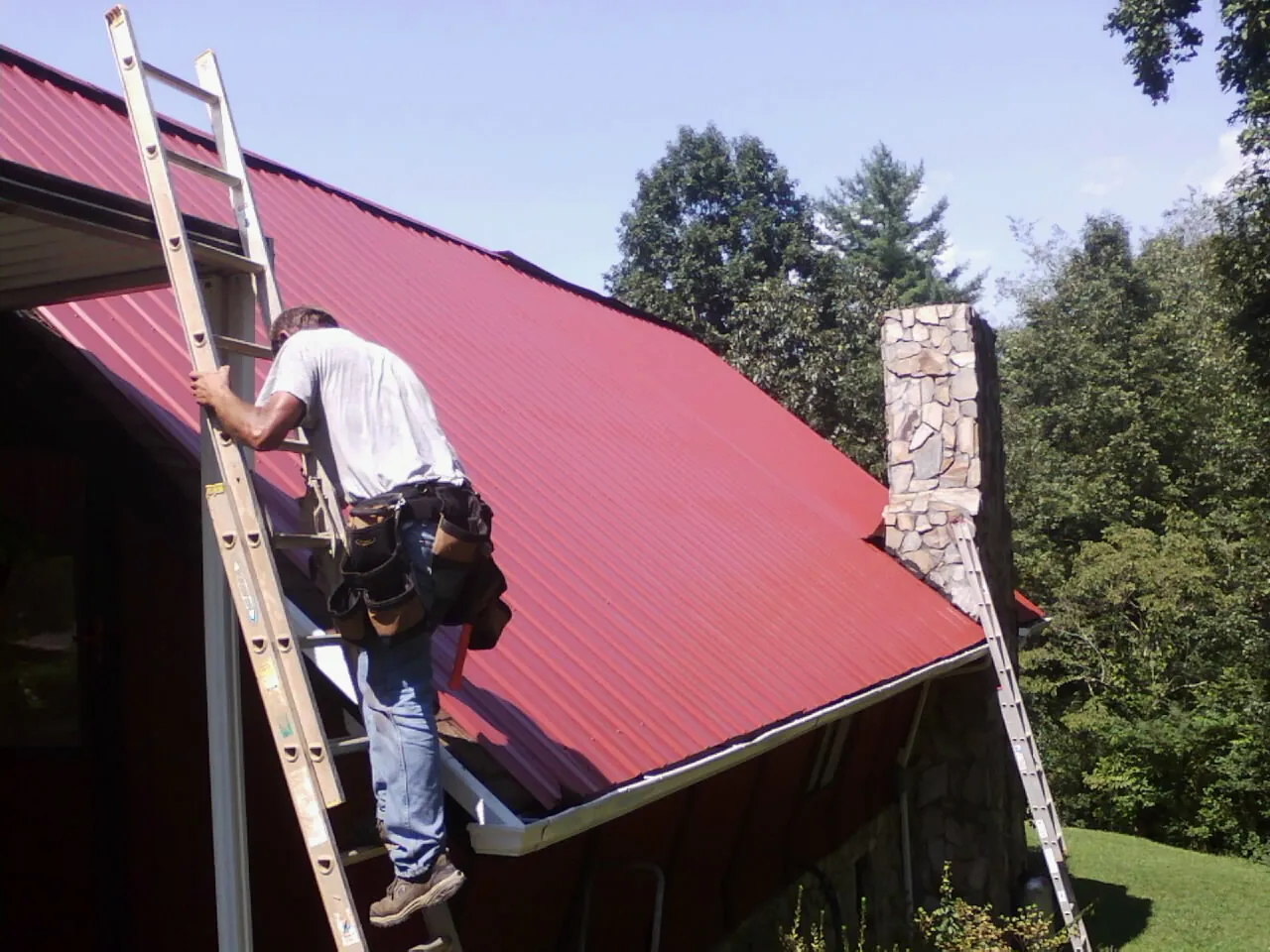 Roofer on a ladder beside a red metal roof and stone chimney — Soffit & Fascia Repair by Roofing Brothers in Richlandtown